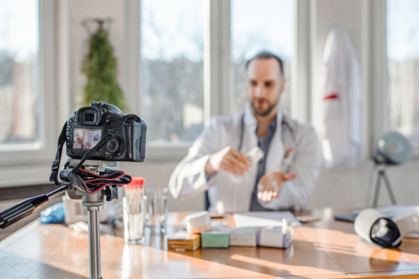 Männlicher Arzt sitzt vor einer Kamera und zeigt Medikament